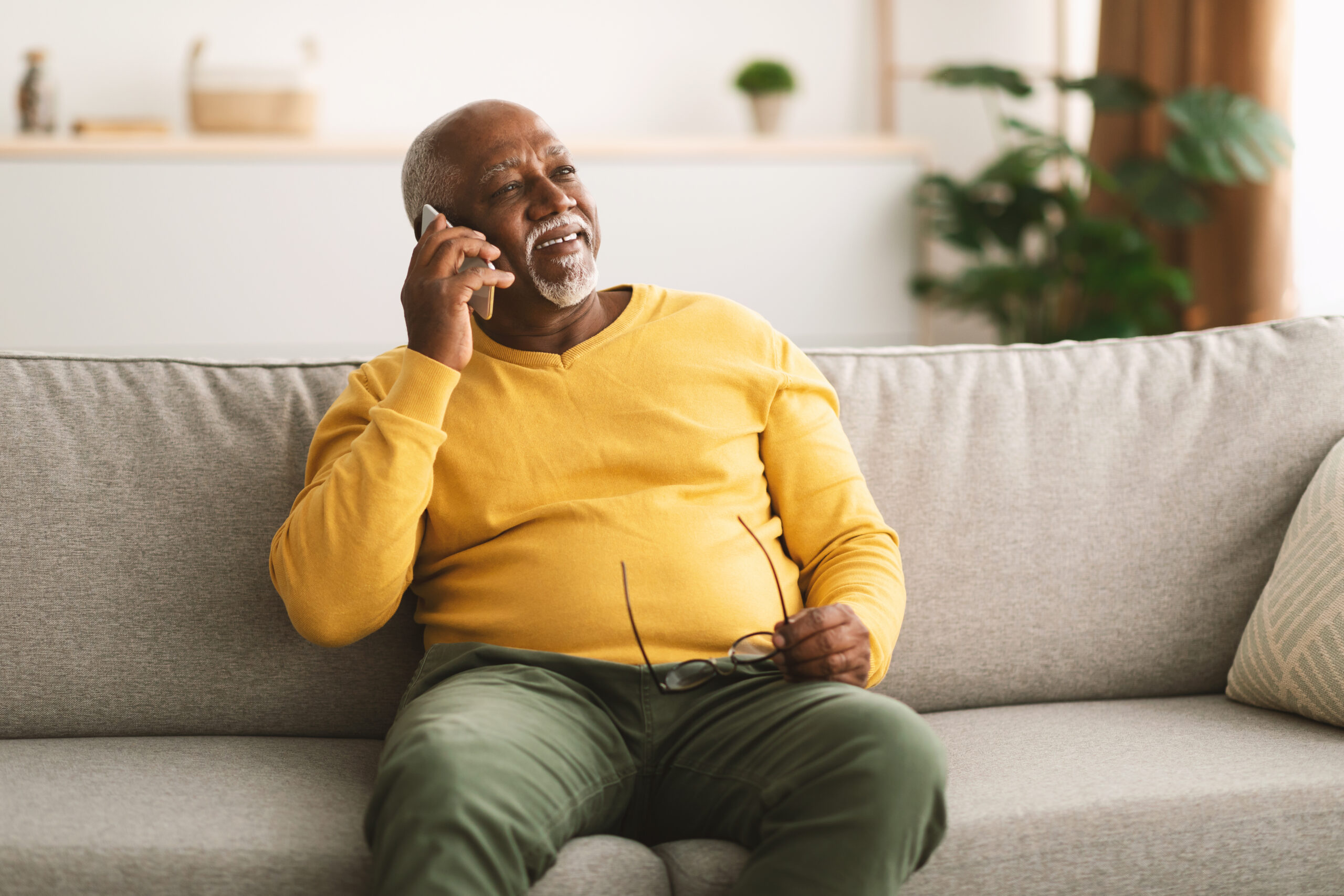 Senior African American Man Talking On Cellphone Having Pleasant Phone Conversation Sitting On Sofa At Home, Looking Aside And Smiling.