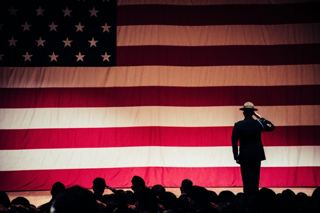 Wide shot of a man standing on a stage and facing and American flag, backlit and saluting. A crowd of people sit in front of the stage, also backlit.