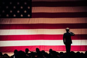 Wide shot of a man standing on a stage and facing and American flag, backlit and saluting. A crowd of people sit in front of the stage, also backlit.