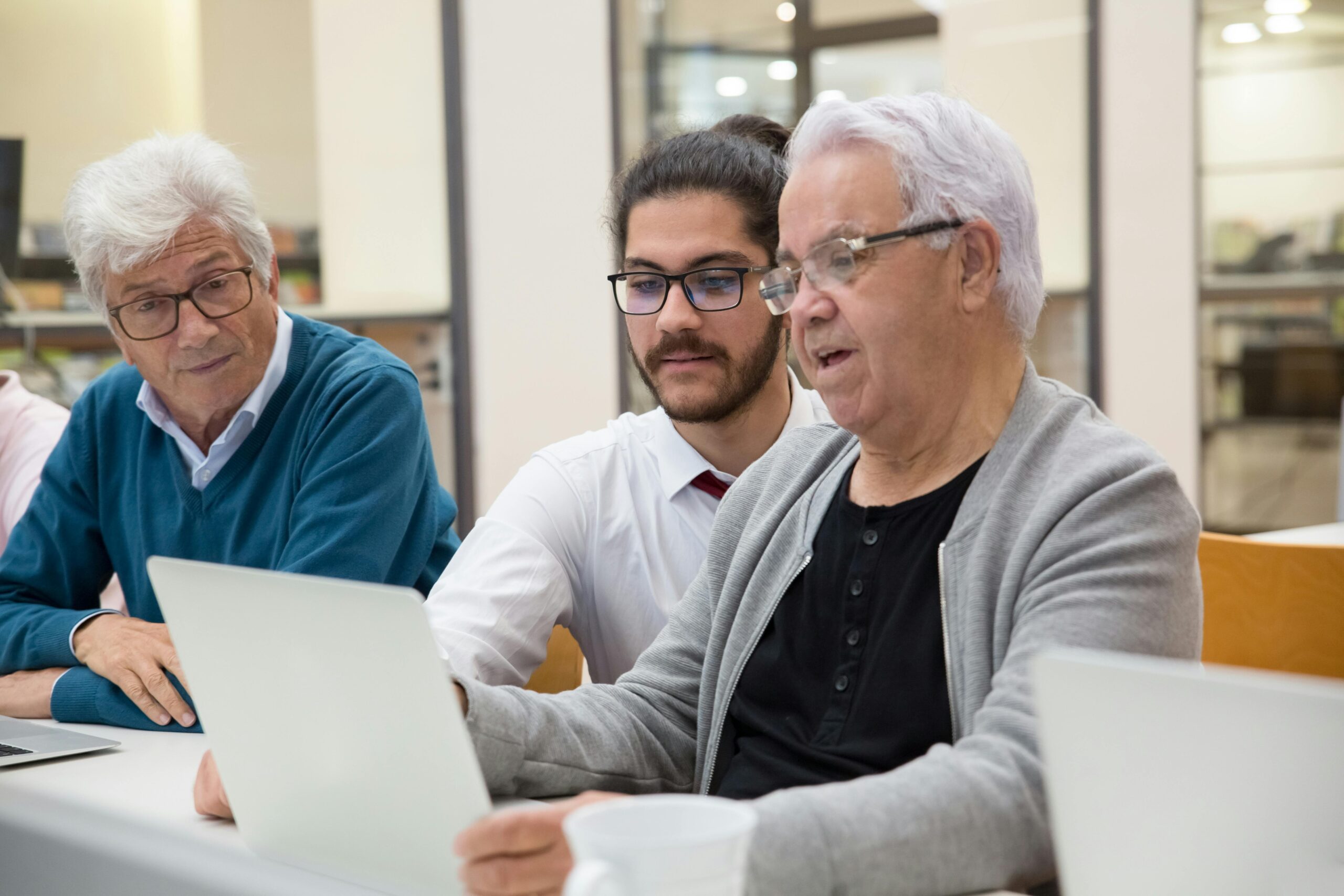 A young man sits between two elderly men using laptops, pointing to the screen.