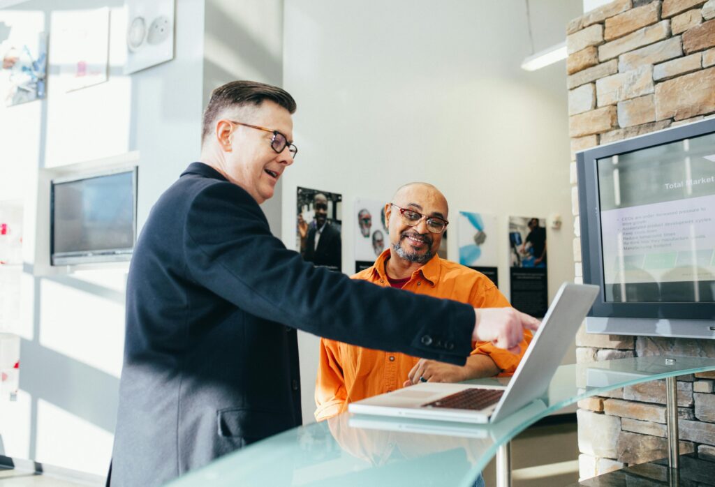 Two men in the lobby of an office, looking at a laptop. One mans points at the screen and the other smiles.