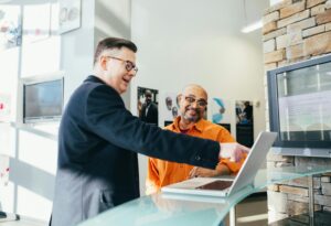 Two men in the lobby of an office, looking at a laptop. One mans points at the screen and the other smiles.