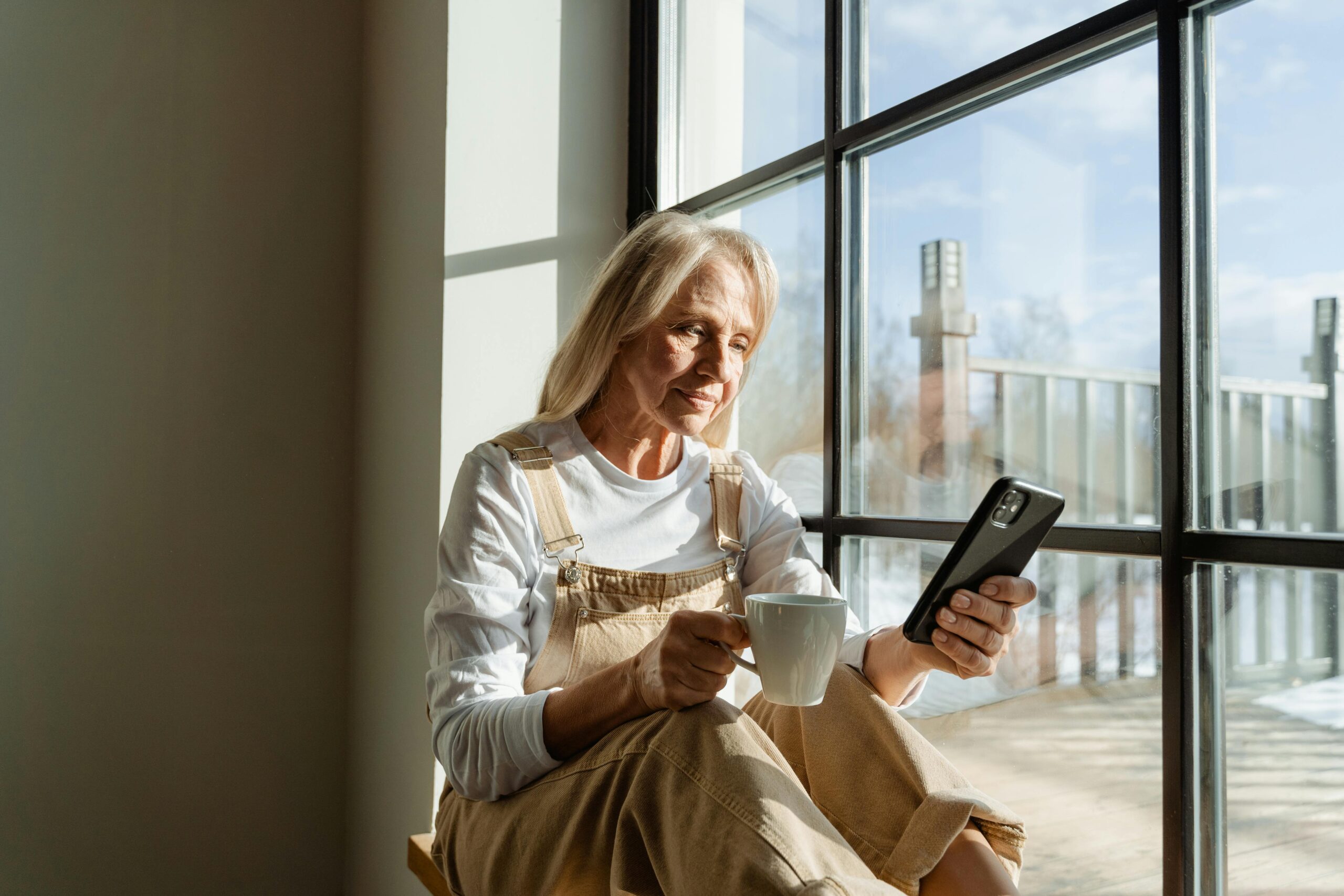 An elderly woman in overalls and a long-sleeve shirt sits on a window bench, holding a mug in one hand and a phone in the other.