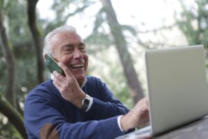 Elderly man using a laptop and holding a cell phone to his ear while smiling.