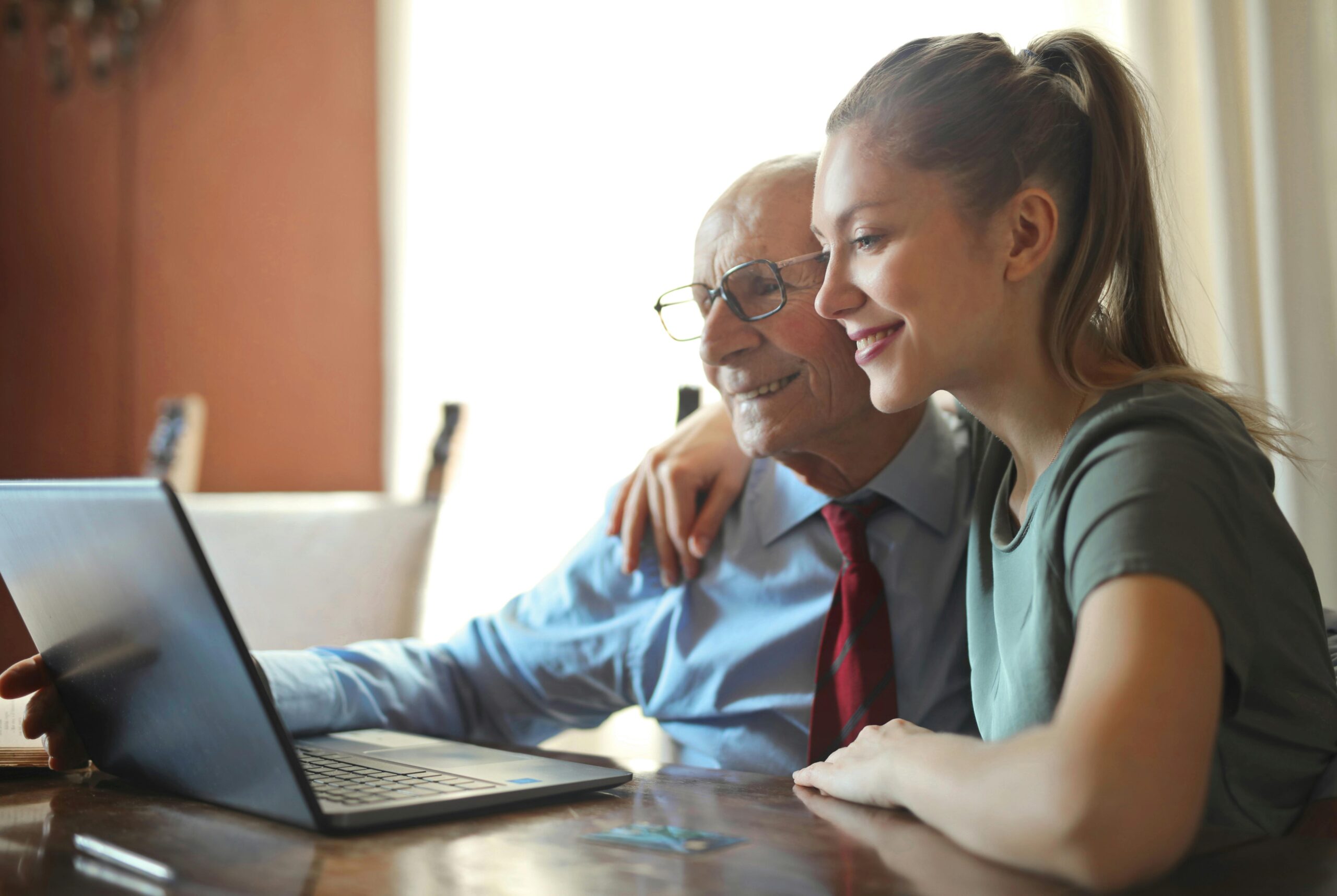 Young woman and elderly man smiling and looking at a laptop. They are sitting and the young woman has her arm around the man's shoulders.
