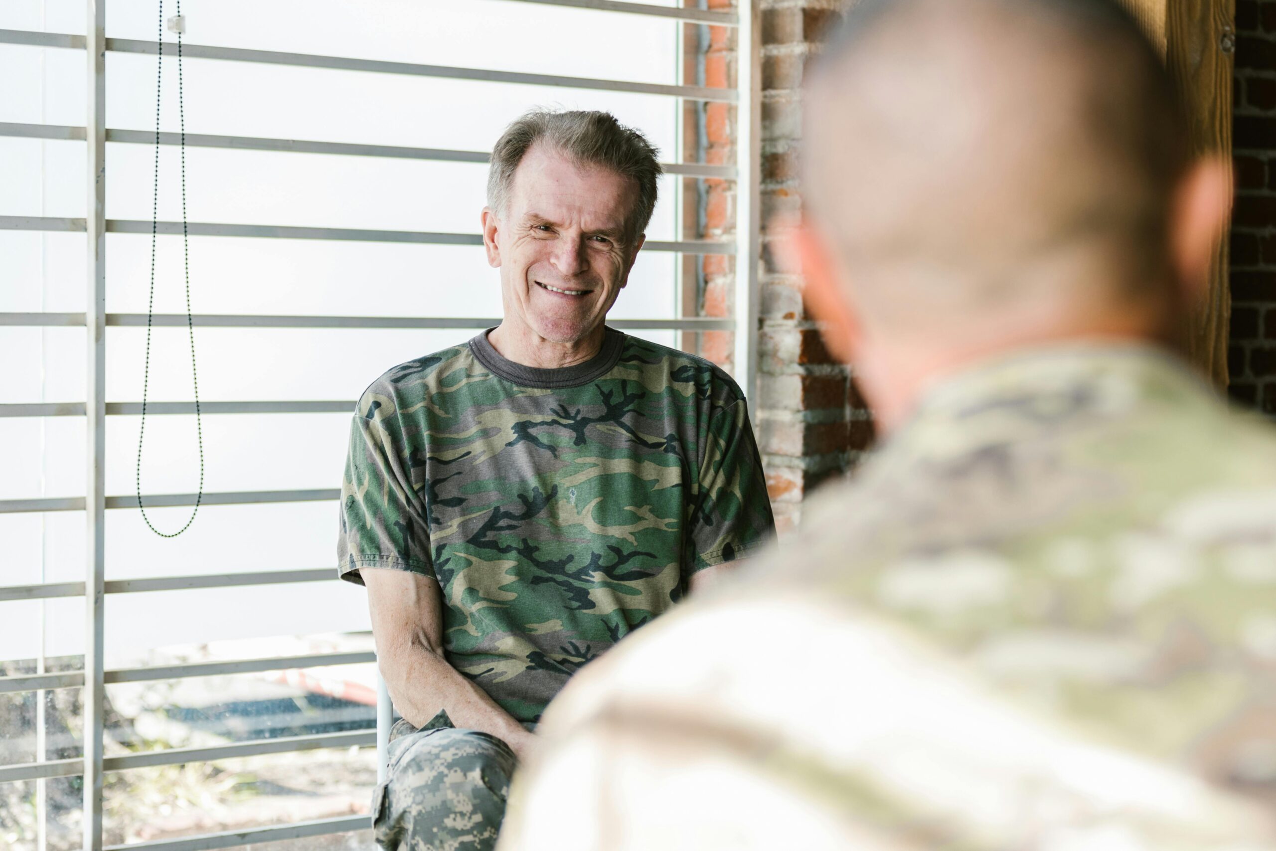 Over-the-shoulder shot of a man in fatigues facing an elderly man in a camouflage t-shirt and army pants.