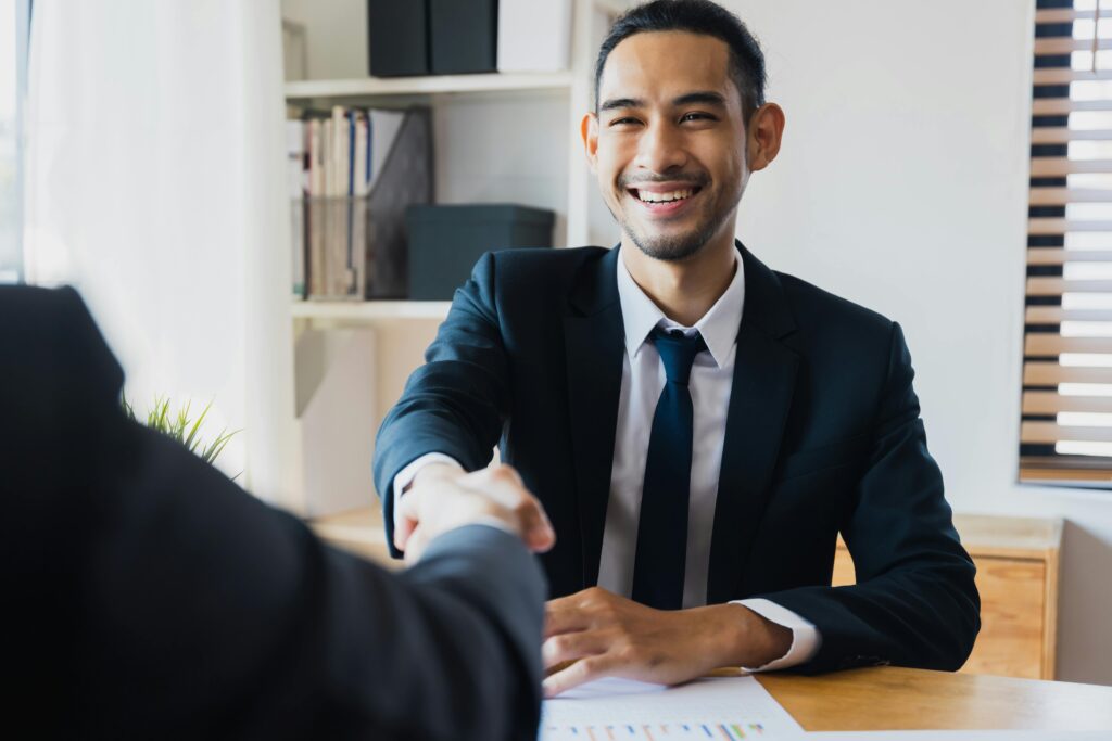 Over-the-shoulder shot of a person in a suit shaking hands with a young businessman in a suit. They are seated at a table in an office.