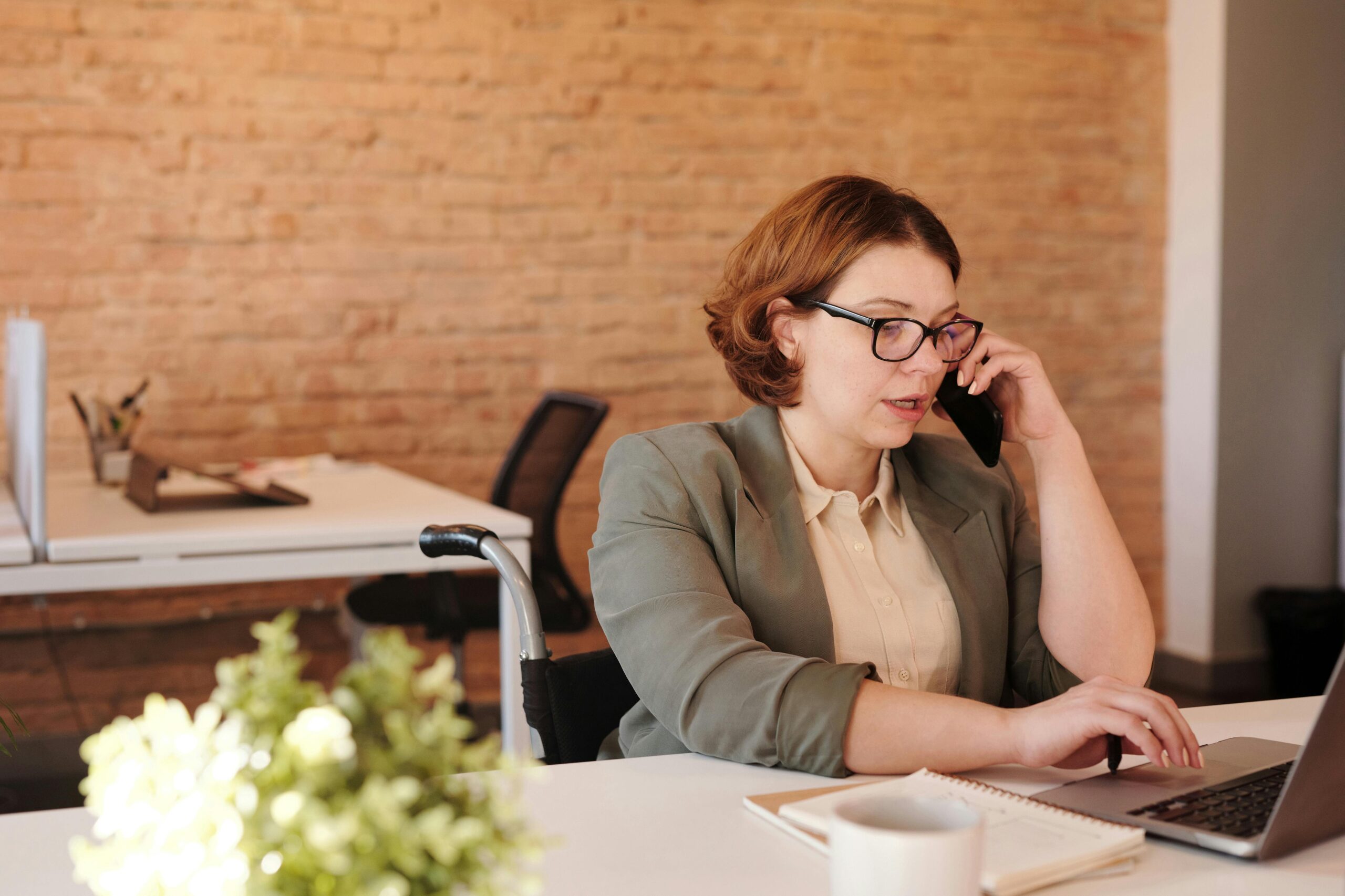 Insurance agent in a wheelchair working at a desk, speaking on the phone while using a laptop to follow up with clients in a modern office.
