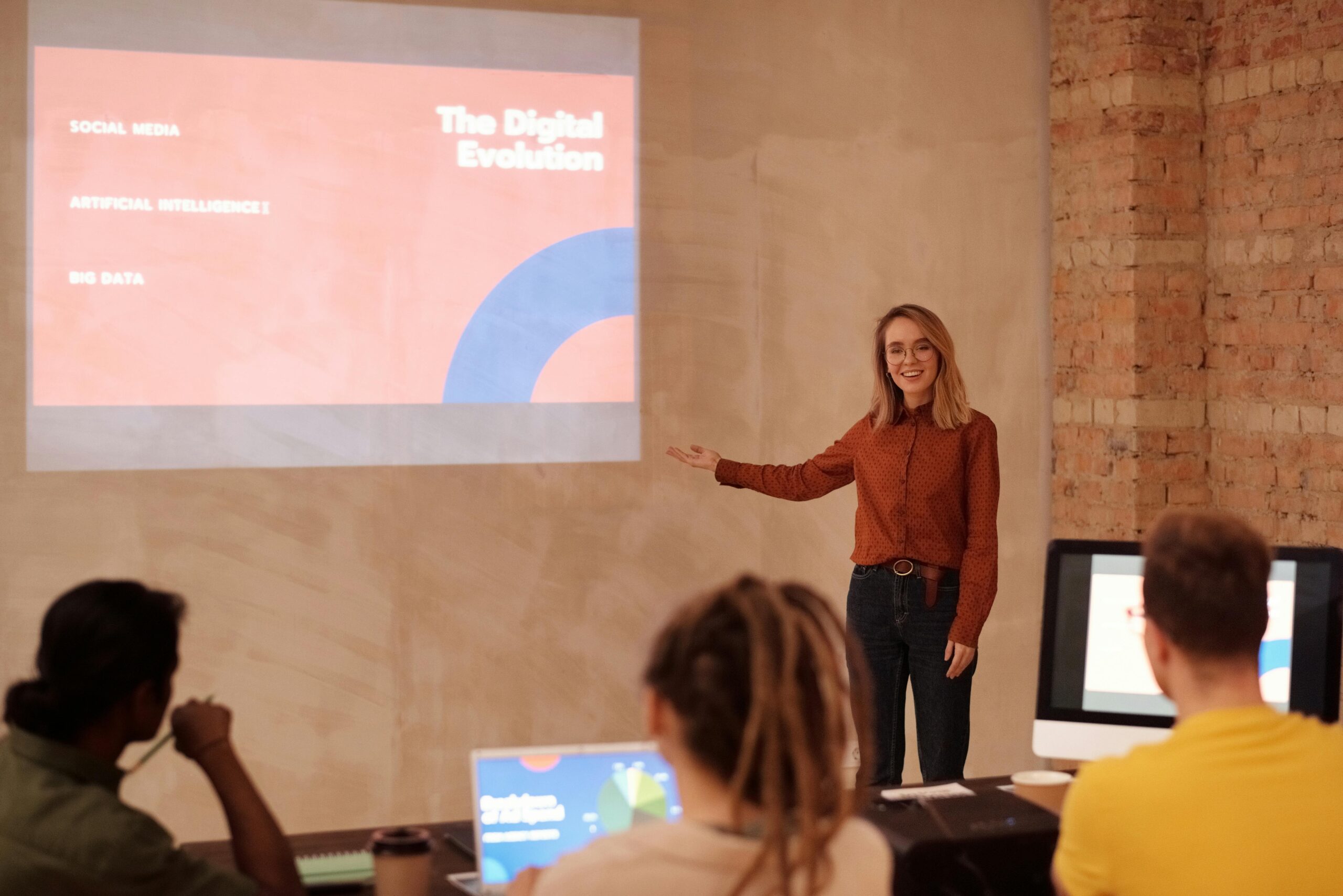 A woman in professional clothing stands at the front of a classroom. She gestures to a slide projected on the wall that says "The Digital Evolution: Social Media, Artificial Intelligence, Big Data"