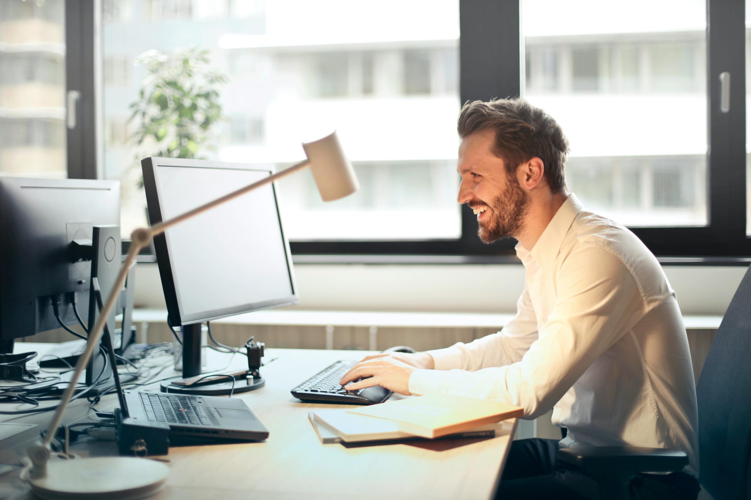 A man smiles while typing, sitting at a desk that holds a laptop, desktop, lamp, and a notepad.