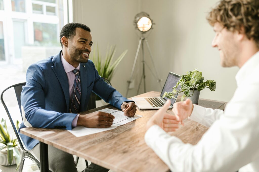 An insurance agent in a blue suit smiles while meeting with a Gen X client across a desk. Papers and a laptop are on the table.