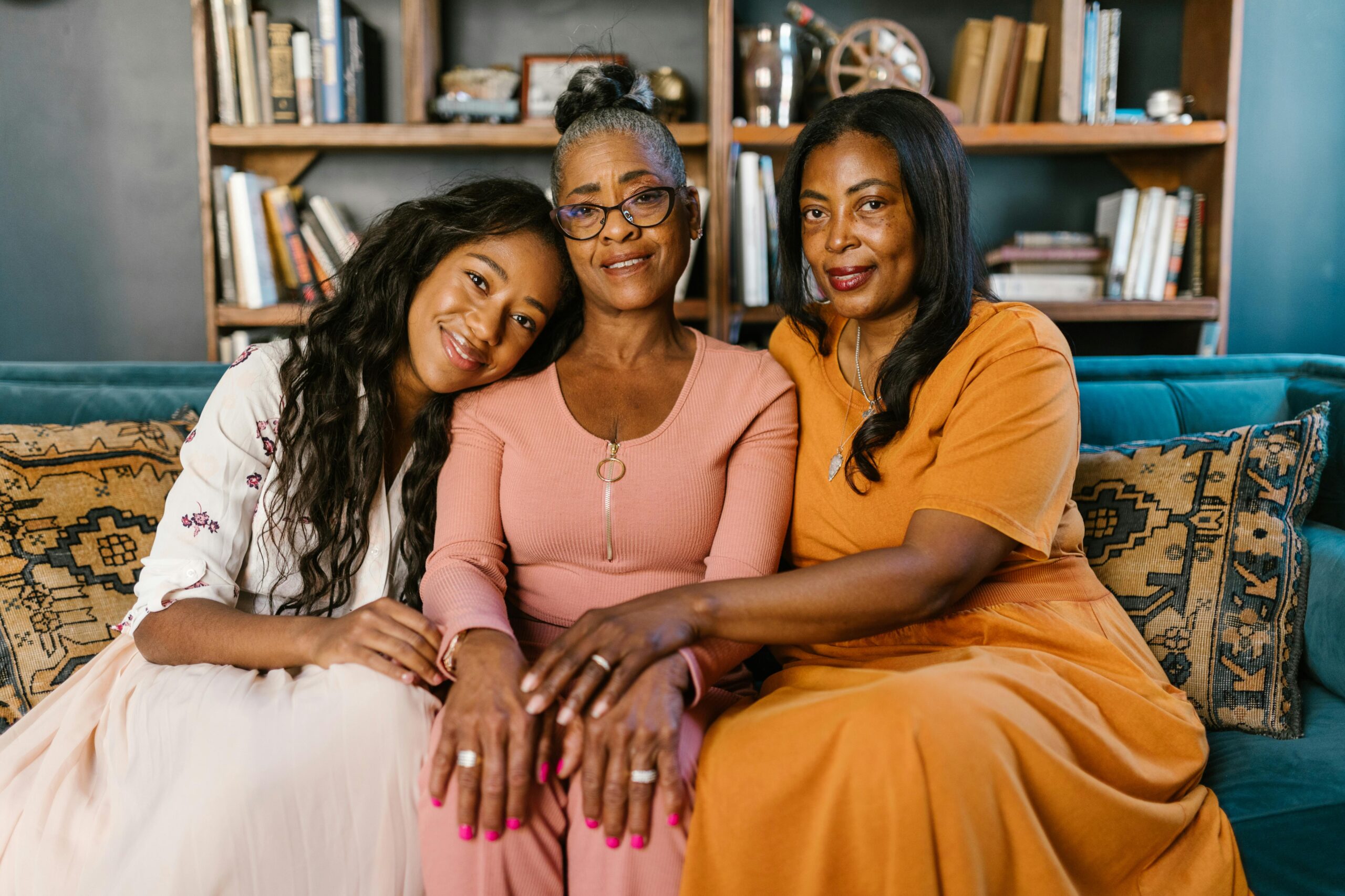 Three generations of women, Boomer, Gen X, and Gen Z, sit closely together on a couch at home. The younger woman rests her head on her grandmother’s shoulder while the Gen X mother smiles beside them.