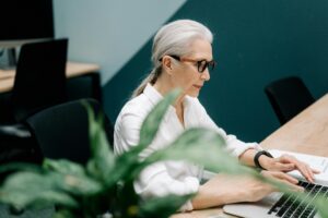 An older woman wearing glasses types on a laptop in an office space with a plant taking up the foreground.