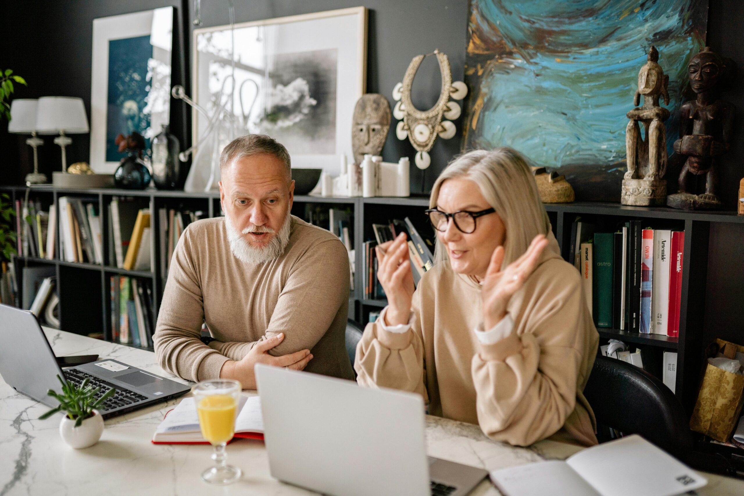 An older couple sits in front of their individual laptops in a cluttered office.